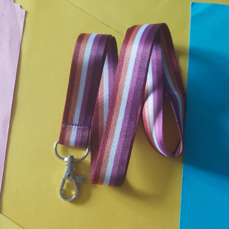 A pink, orange and white striped fabric lanyard with a silver metal lobster clasp, laid out on a yellow background with colourful paper edges visible.