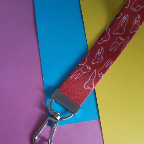 A close-up of a red fabric lanyard strap with a white teeth pattern, attached to a silver metal swivel clasp, laid diagonally over colourful paper backgrounds.