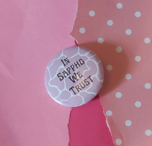 A round pinback button badge with the text 'IN SAPPHO WE TRUST' in black lettering, set against a background of torn pink paper and pink polka-dot paper.