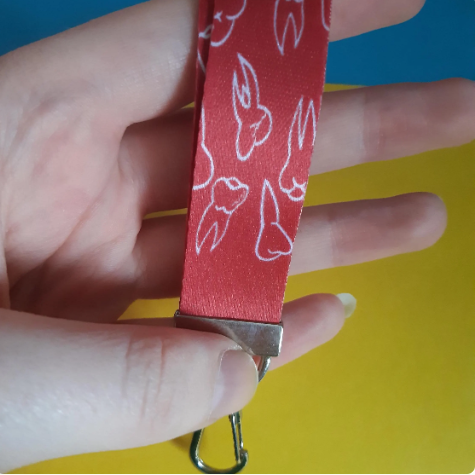 A close-up of a hand holding a red fabric keychain strap with white tooth outlines and a silver metal clasp against a bright yellow and blue background.