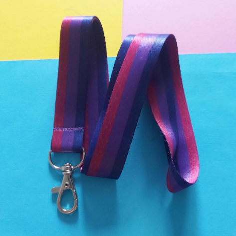 A cotton lanyard with alternating purple, blue and magenta stripes, featuring a silver metal lobster clasp, laid out on a colourful background of yellow, pink, and blue paper.